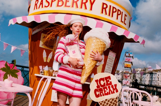 A person in pink holds an oversized ice cream and Jo Malone London cologne on a pier, with pink seating, and a giant cone hut.